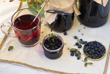 On a light background, on a beautiful fabric napkin, there are several packaged glass jars with blueberry jam, and in front there is a fresh tasty blueberry with green leaves on the branches and a swe
