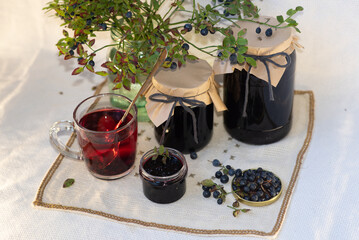 On a light background, on a beautiful fabric napkin, there are several packaged glass jars with blueberry jam, and in front there is a fresh tasty blueberry with green leaves on the branches and a swe