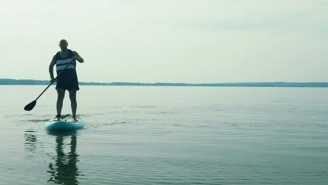 A man with a backpack on a SUP board swims in the lake in the evening against the background of a sky with clouds. Overall plan