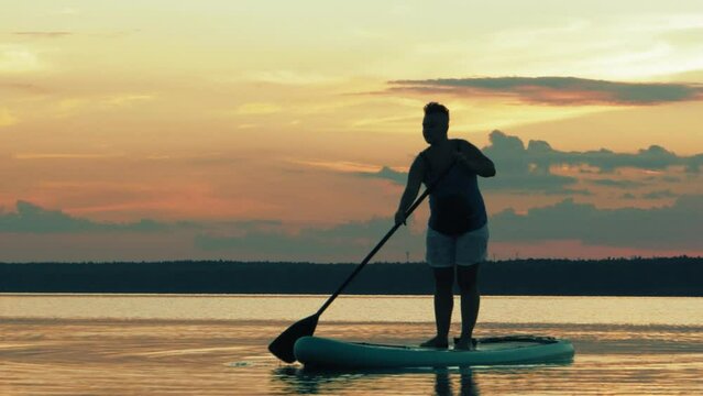 A woman in shorts on a sup board swims in the lake against the backdrop of a golden sunset in the evening. Overall plan