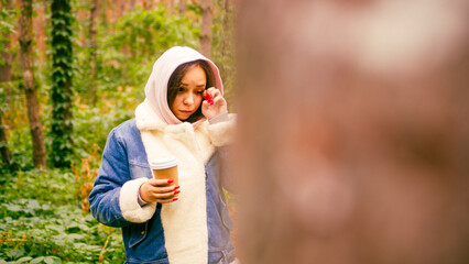 Fototapeta premium Young woman rubs her eye, holding paper cup with coffee, standing in forest.