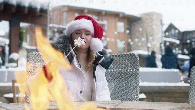 Hand of caucasian woman with marshmallow on a stick roasting over a bonfire on winter day. Cozy fireplace in modern luxury ski resort village in Aspen. Woman in red Santa hat on holiday vacation in US