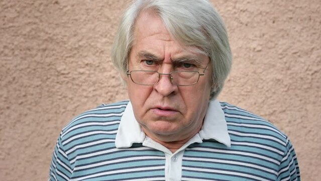 Closeup portrait of an old man, grandfather looking very unhappy, mad and angry isolated on concrete wall background. Senior man facial expression. Menacing Man with gray hair. Negative elderly person