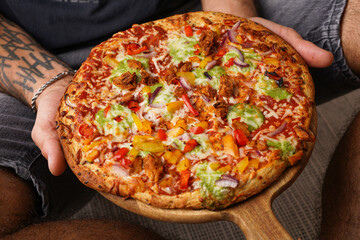 Male sitting and with hands with tattoos holding a vegetarian clean pizza with quinoa crust, basil sauce and colorful vegetables