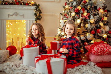 Obraz na płótnie Canvas Two adorable girls in checkered dresses sitting near Christmas tree and opening their presents