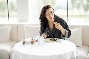 Beautiful woman in bathrobe enjoys delicious meal, sitting in white restaurant of haught cuisine during vacations at luxury spa resort