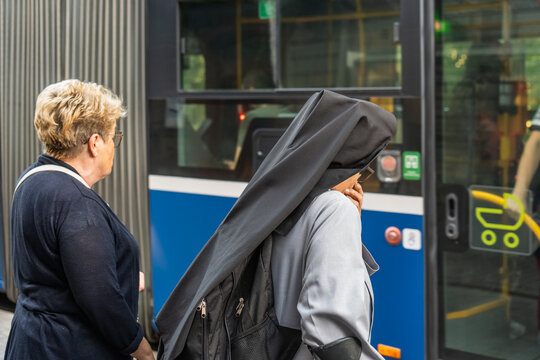 Catholic nun in monastic clothes stands thoughtfully at the door of a city bus at a public transport stop in Krakow, an ordinary woman behind a nun in line to enter the bus, two women