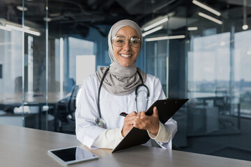 Portrait of successful muslim female doctor in hijab, arabic woman working in modern clinic office, smiling and looking at camera, female doctor in glasses and white medical coat with stethoscope.