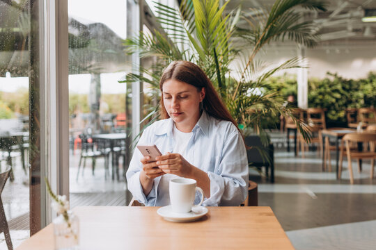 White brunete woman using smartphone while sitting in cafe indoors. Young woman at cafe drinking coffee or tea indoors on a rainy day. Concept of easy breakfast.