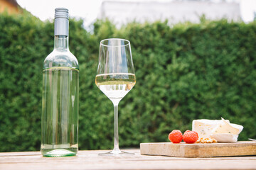 A bottle of white wine with a glass stands on a wooden table next to cheese and fresh berries against the background of a green hedge.