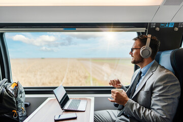 Handsome businessman is having a good time while traveling by high-speed train. He is using laptop computer and wireless headphones for online communication, gaming and entertainment.