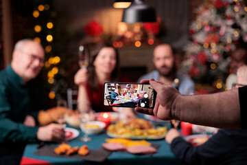 Family christmas celebration photo on smartphone screen, selective focus, people looking at mobile phone camera, blurred background. Xmas holidays, friends gathering together