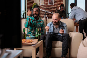 Fototapeta premium Cheerful people celebrating video games win at house gathering, using tv console to play competition. Men feeling happy about winning, having fun wiht friends at party with beer.