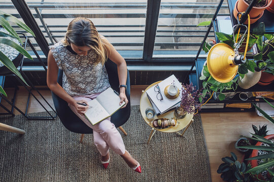 Overhead View Of Businesswoman Writing In Diary While Sitting On Chair At Home Office