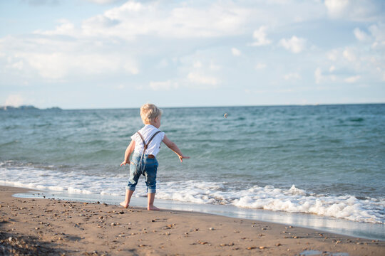 Rear view of baby boy skimming stone while standing at beach against cloudy sky