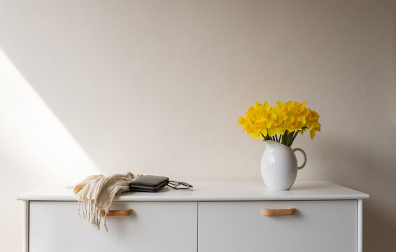 Yellow daffodils in white jug on side table next to scarf, purse and glasses against beige wall with sunlight (selective focus)