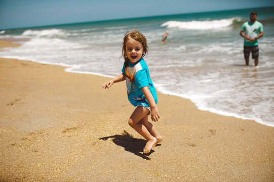 Happy daughter playing at beach while father standing on shore during sunny day