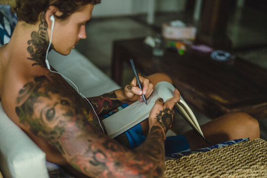 Side view of shirtless tattooed man listening music while sketching on notebook at home