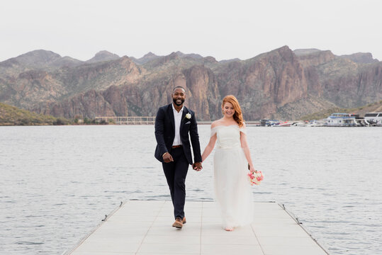 Full length of newlywed couple holding hands while walking on pier over lake against mountains