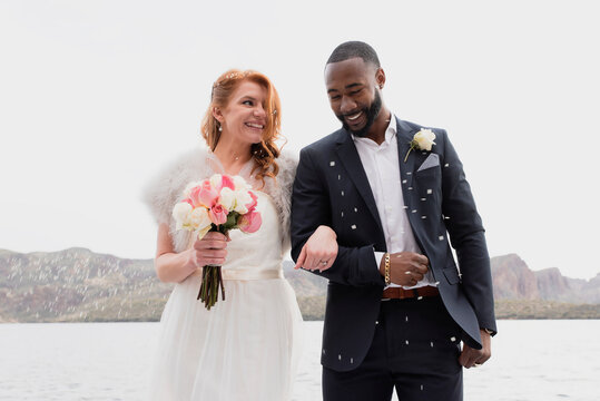 Happy newlywed couple with bouquet standing by lake against clear sky
