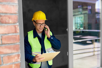 construction business and building concept - happy smiling male builder in helmet and safety west with clipboard calling on smartphone at window