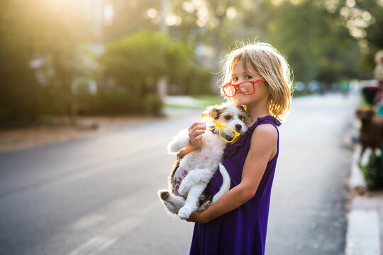 Side view portrait of playful girl with eyeglasses carrying dog while standing on road during sunset