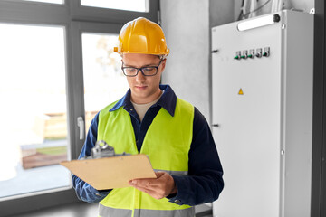 construction business and building concept - male electrician or worker in helmet and safety west with papers on clipboard and pencil at electric board