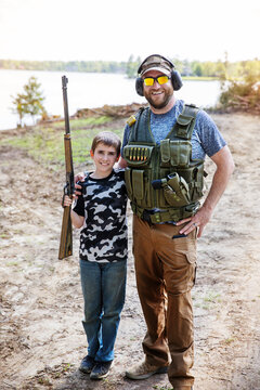 Portrait of son holding rifle while standing with father on field against sky