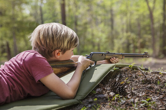 Boy aiming rifle while lying on mat in forest