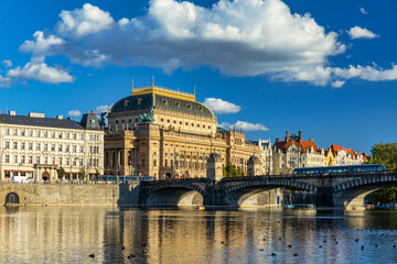 Fototapeta premium National Theatre located in Prague, Czech Republic on the Vltava River. Legion bridge on river Vltava. National Theatre (Narodni Divadlo) on embankment in sunbeams of evening sunset sunlight. Prague