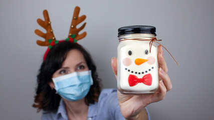 Woman in a mask and reindeer headband showing snowman. Can with a Christmas face. Medical mask,...