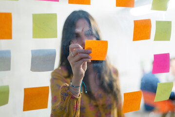 Businesswoman sticking colorful adhesive notes on window in creative office seen through glass