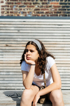 Thoughtful teenage girl with hand on chin looking away while sitting against wall in city