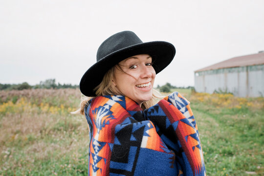 Portrait of happy woman with blanket wearing hat while standing on grassy field against clear sky