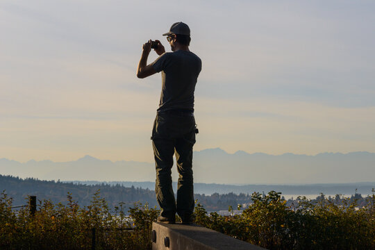 Rear view of man photographing landscape with mobile phone while standing on retaining wall against cloudy sky during sunset