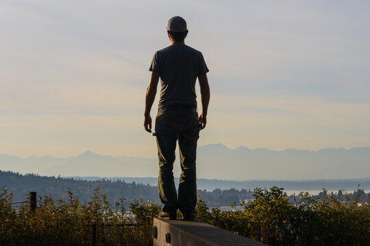 Rear view of man looking at view while standing on retaining wall against cloudy sky during sunset