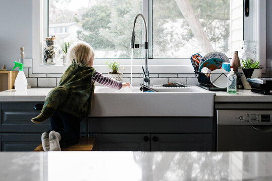 Rear view of girl washing hands in kitchen sink while kneeling on stool at home