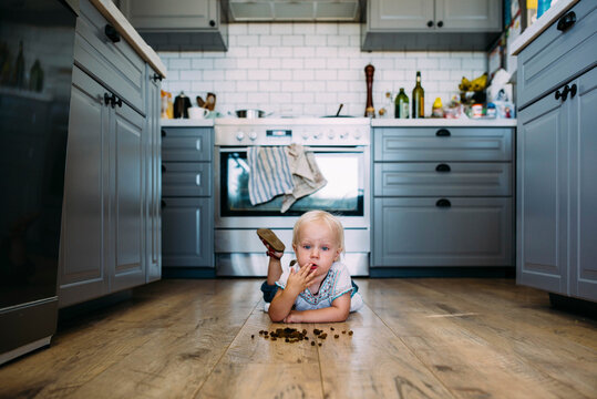 Cute thoughtful girl eating food while lying on hardwood floor in kitchen at home