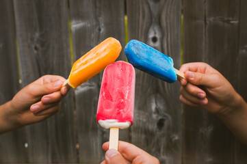 Cropped hands of brothers holding colorful flavored ice by fence at backyard