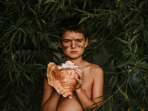 Portrait of shirtless boy holding conch shell while standing amidst plants