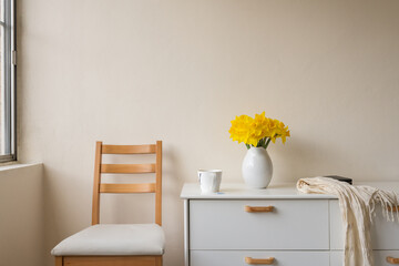 Yellow daffodils in white jug on side table next to chair against beige wall (selective focus)