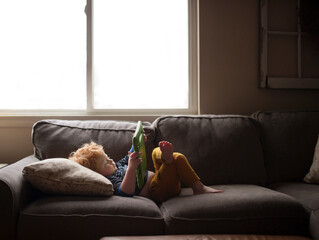 Young boy lays on couch at home watching his tablet by window