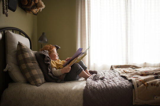 Young boy sits on bed looking through a book at home next to window