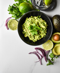 Close up of bowl of guacamole and it's ingredients on a white counter.