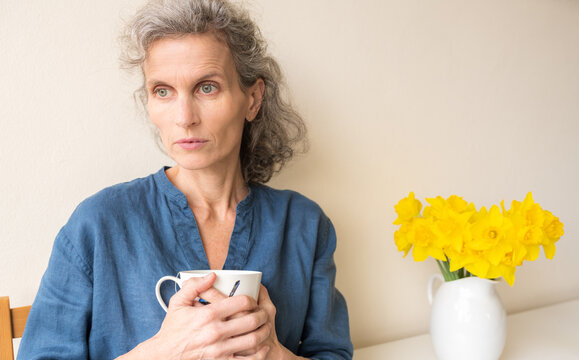 Middle aged woman with grey wavy hair and blue top seated with cup next to daffodils, looking pensive (selective focus)