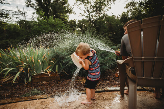 Toddler boy dumping bucket of water while dad waters garden