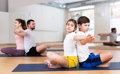 Fototapeta premium Tween girl with brother sitting on mat in modern yoga studio during family workout, doing exercises in pair ..