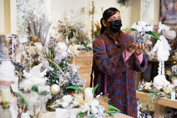 Latino american woman in face mask preparing for Christmas and choosing gifts and decorations at flower shop
