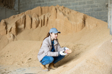 Positive female farm worker holding scoop with soybean hulls, natural cattle feed, at farm storage