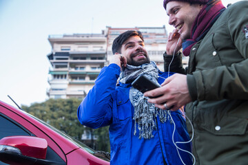 Two happy gay guys smiling while listening music from mobile and sharing headphones next to red car...